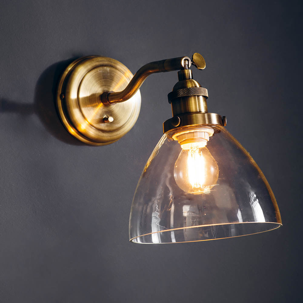 Wentworth Wall Light in brass with adjustable knuckle joint, knurled lamp holder and clear glass dome shade, shown lit against a dark grey wall.