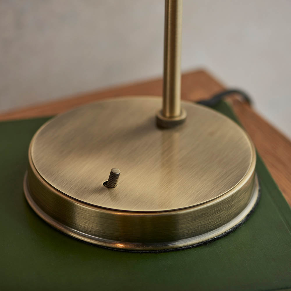 Close-up of the Wentworth Table Lamp's brass circular base with knurled toggle switch, rising rod, and black flex, set on a green surface.