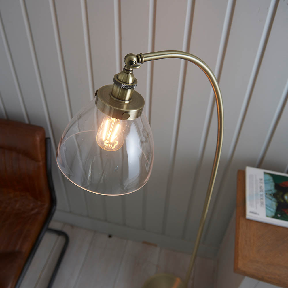 Endon Wentworth floor lamp in brass with adjustable knuckle, knurled lamp holder and clear glass shade lit by a filament bulb, beside a leather chair.