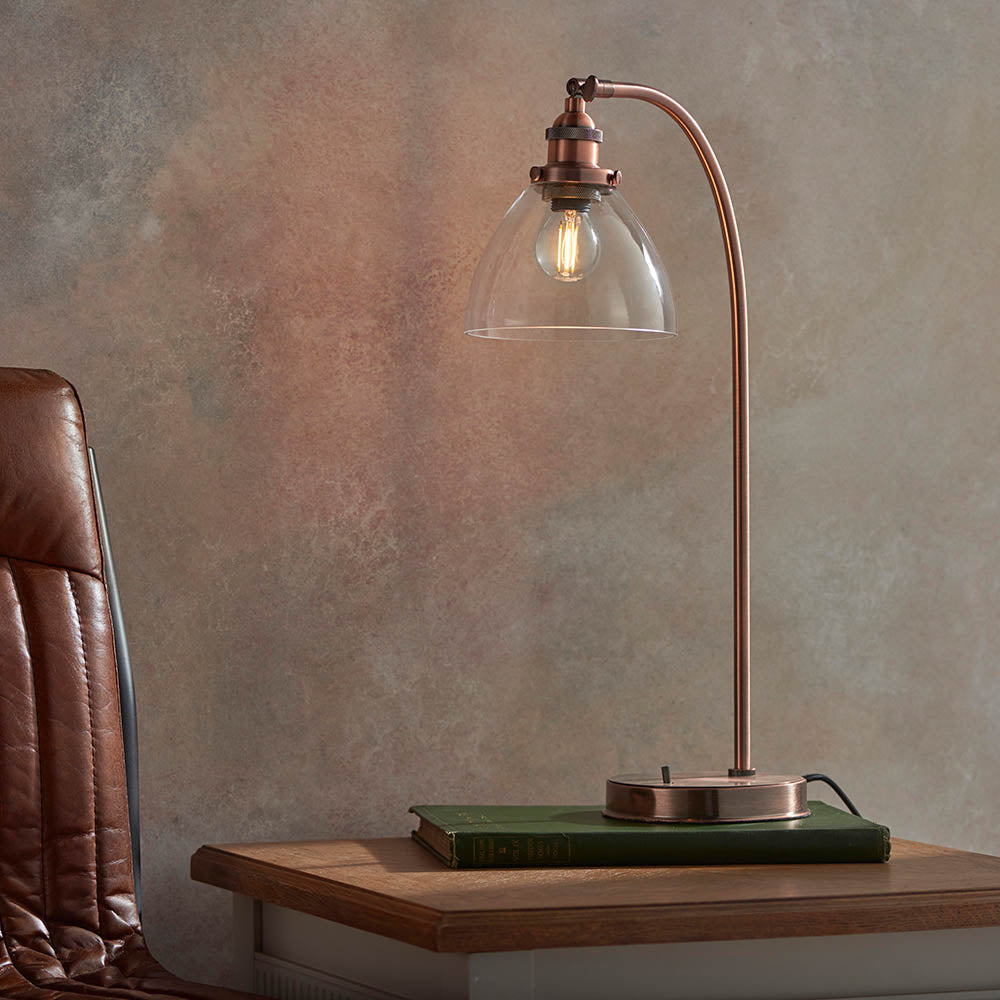 Endon Wentworth copper table lamp with arched arm, clear glass dome shade and filament bulb, set on a green book atop a wooden table beside a leather chair.