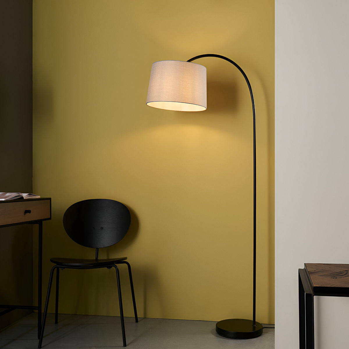 Endon Bevbourne arc floor lamp in matt black with tilting grey fabric shade, beside a black chair against a yellow wall in a living space.