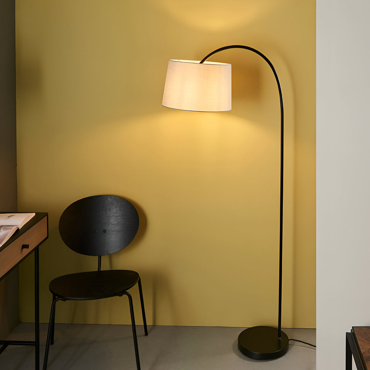 Endon Bevbourne arched floor lamp in matt black with tilting light grey fabric shade, beside a black chair and desk against a yellow wall.