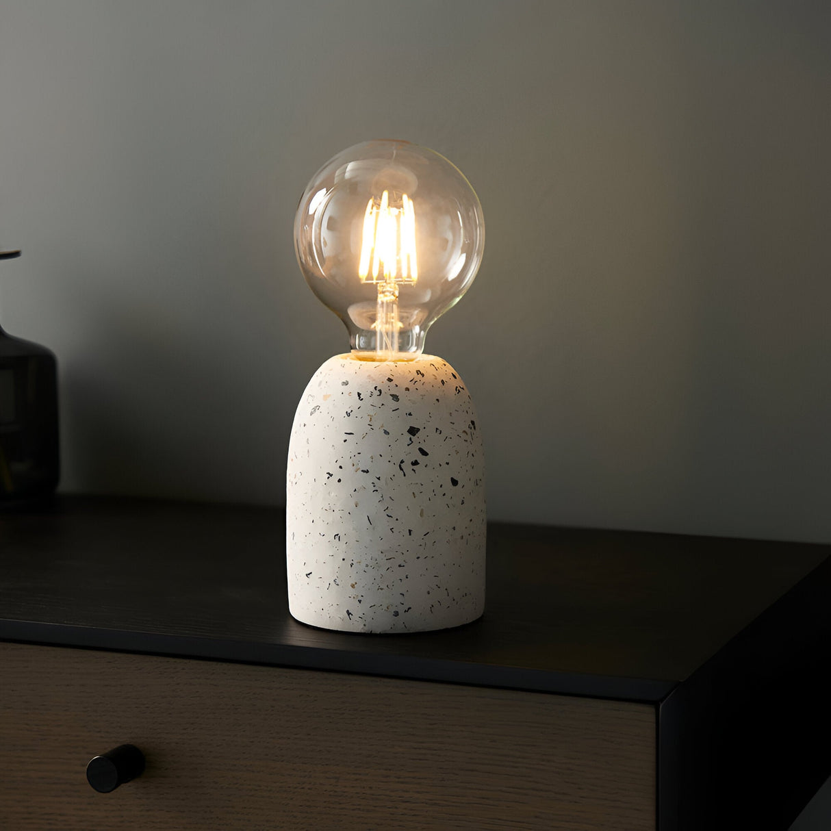 Endon Crangrove small white terrazzo table lamp with speckled matt base and exposed E27 filament globe bulb, lit on a dark wood bedside drawer.
