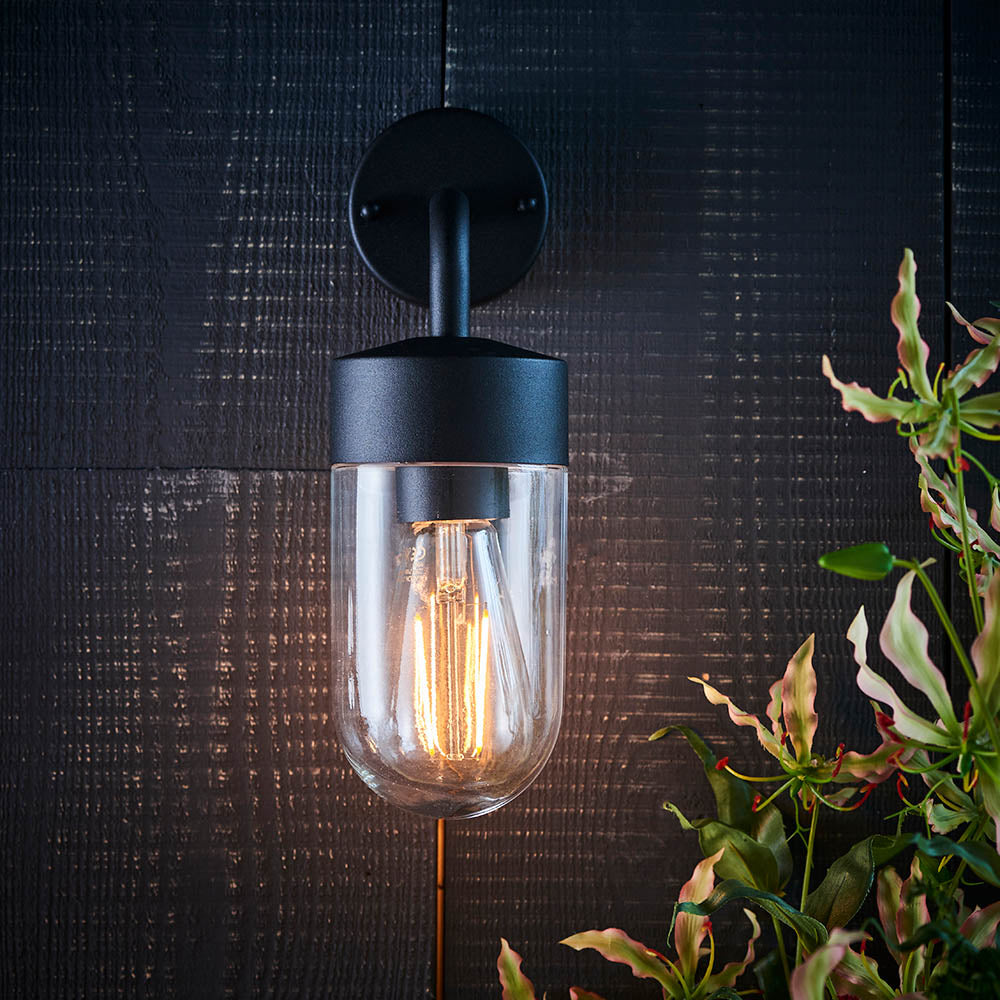 Endon Castfield IP44 outdoor wall light in matt black with a clear glass cylinder shade and exposed E27 filament bulb, mounted on a dark panelled wall.