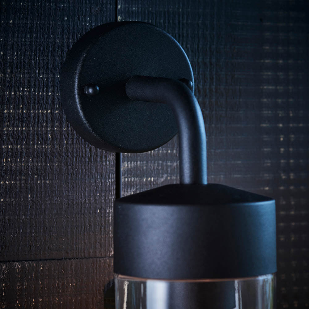 Endon Castfield outdoor wall light in matt black, close-up of the round wall plate and curved arm above the cylindrical shade with clear glass base.