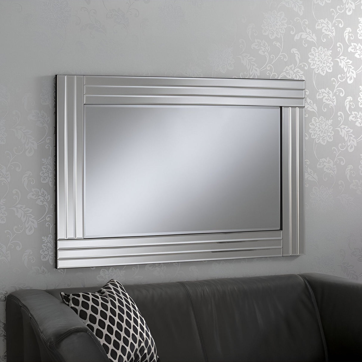 The Edge Rectangle Wall Mirror, featuring a contemporary rectangular design with a thick silver frame, hangs on a light gray wall decorated with floral patterns. Below it, you can spot a dark gray sofa enhanced by a black and white patterned cushion, bringing an element of sophistication to the modern decor.