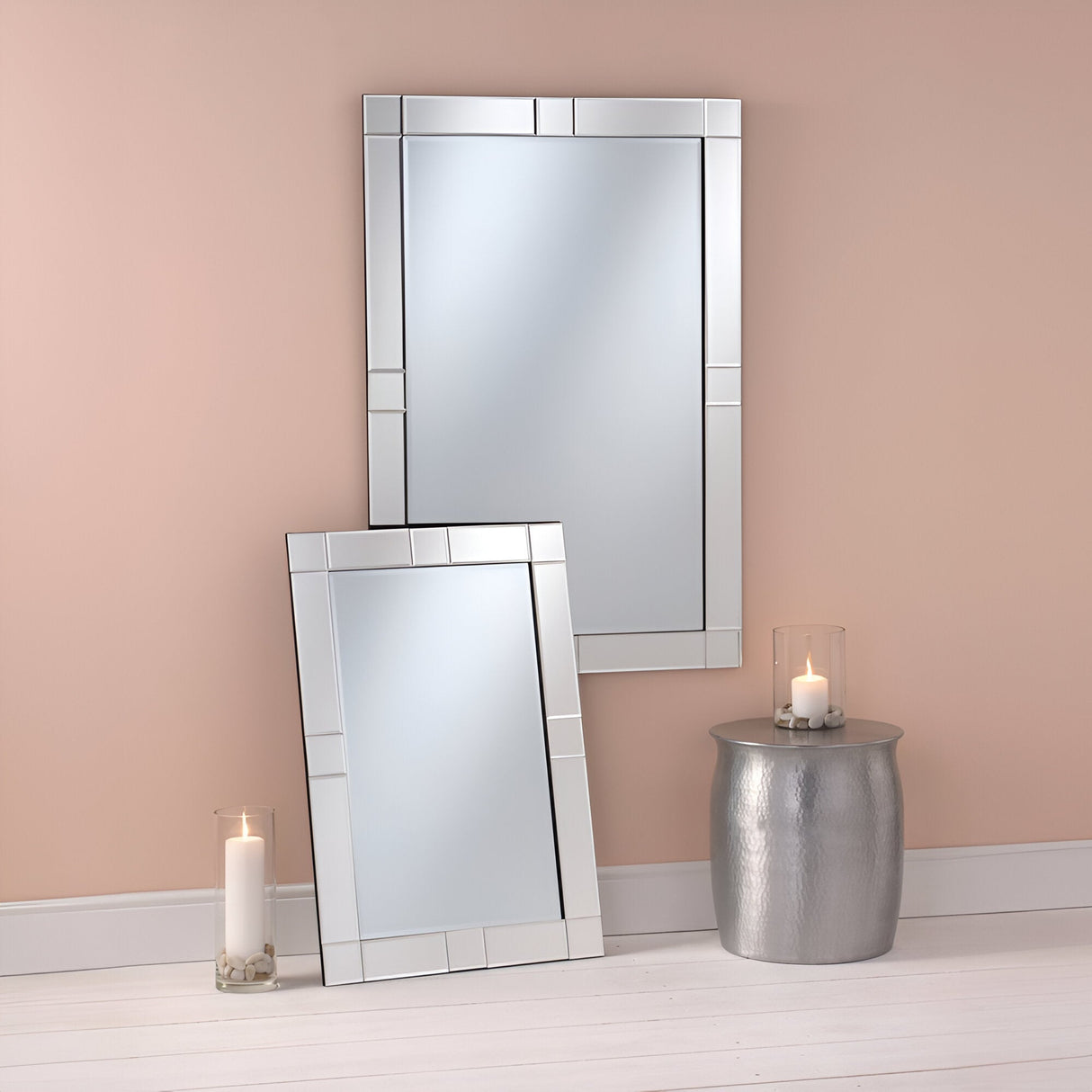 Two Dawn Rectangle Wall Mirrors featuring geometric frames are elegantly positioned against a beige wall—one mirror is resting on the floor, while the other stands upright. Nearby, a silver side table holds a candle in a glass holder, enhancing the minimalist design theme. The versatile decor scene is completed by light wood flooring.