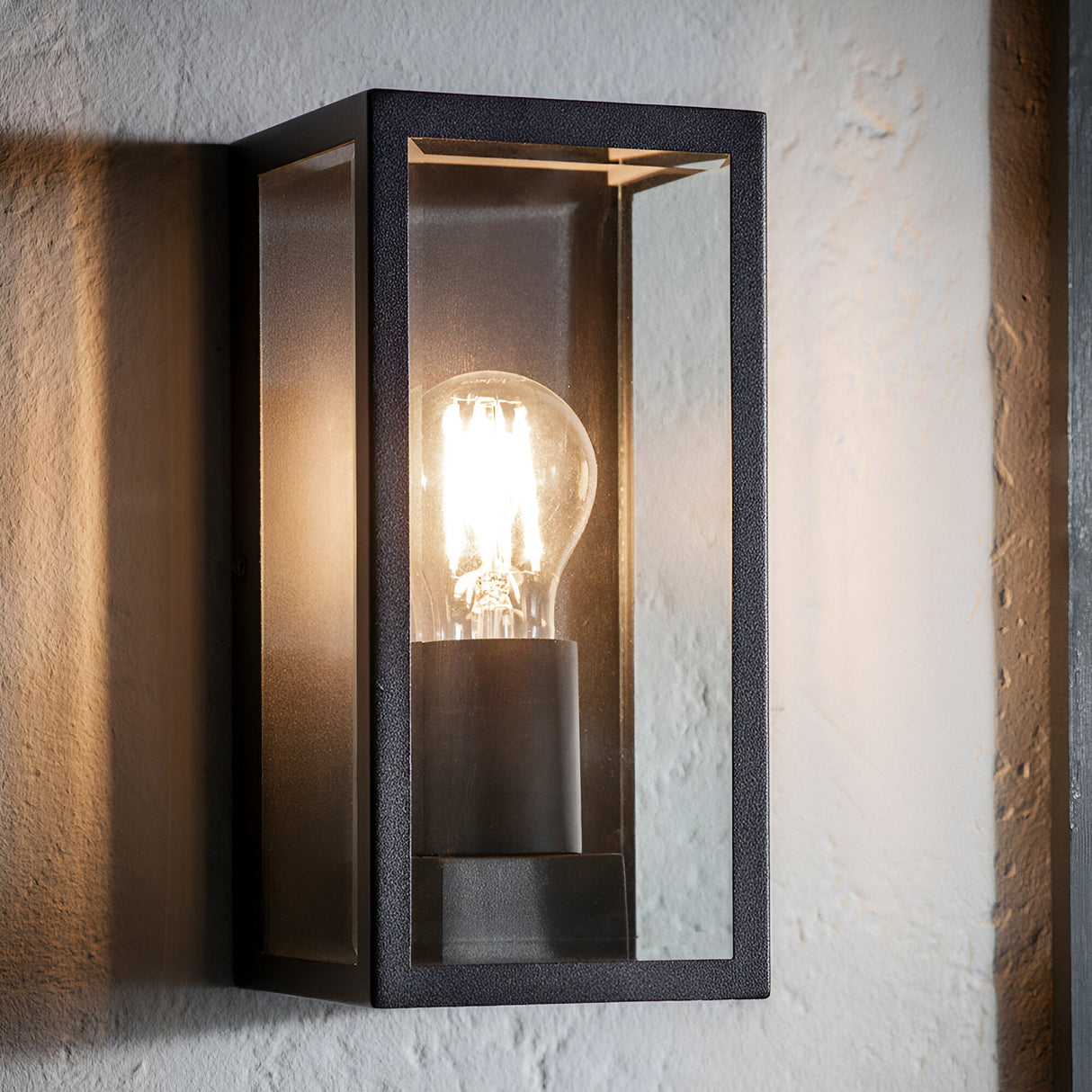 Endon Castwick box lantern wall light in matt black with clear bevelled glass panels and an exposed E27 filament bulb, mounted on a textured pale wall.
