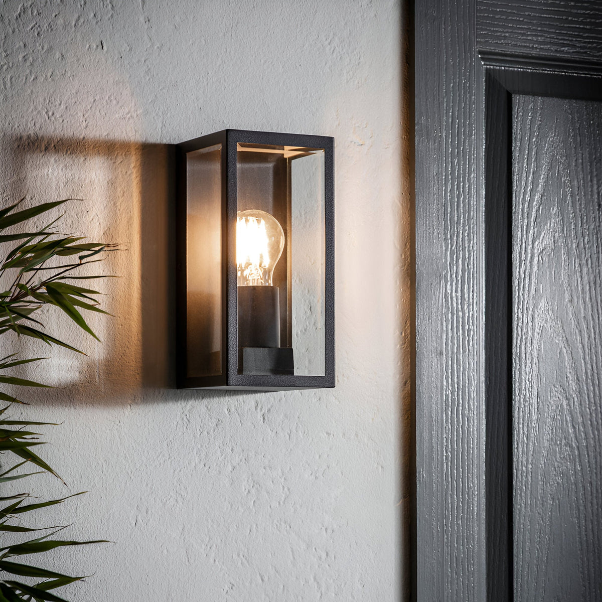 Endon Castwick box lantern wall light in matt black with clear bevelled glass panels and E27 filament bulb, mounted beside a dark wooden door.