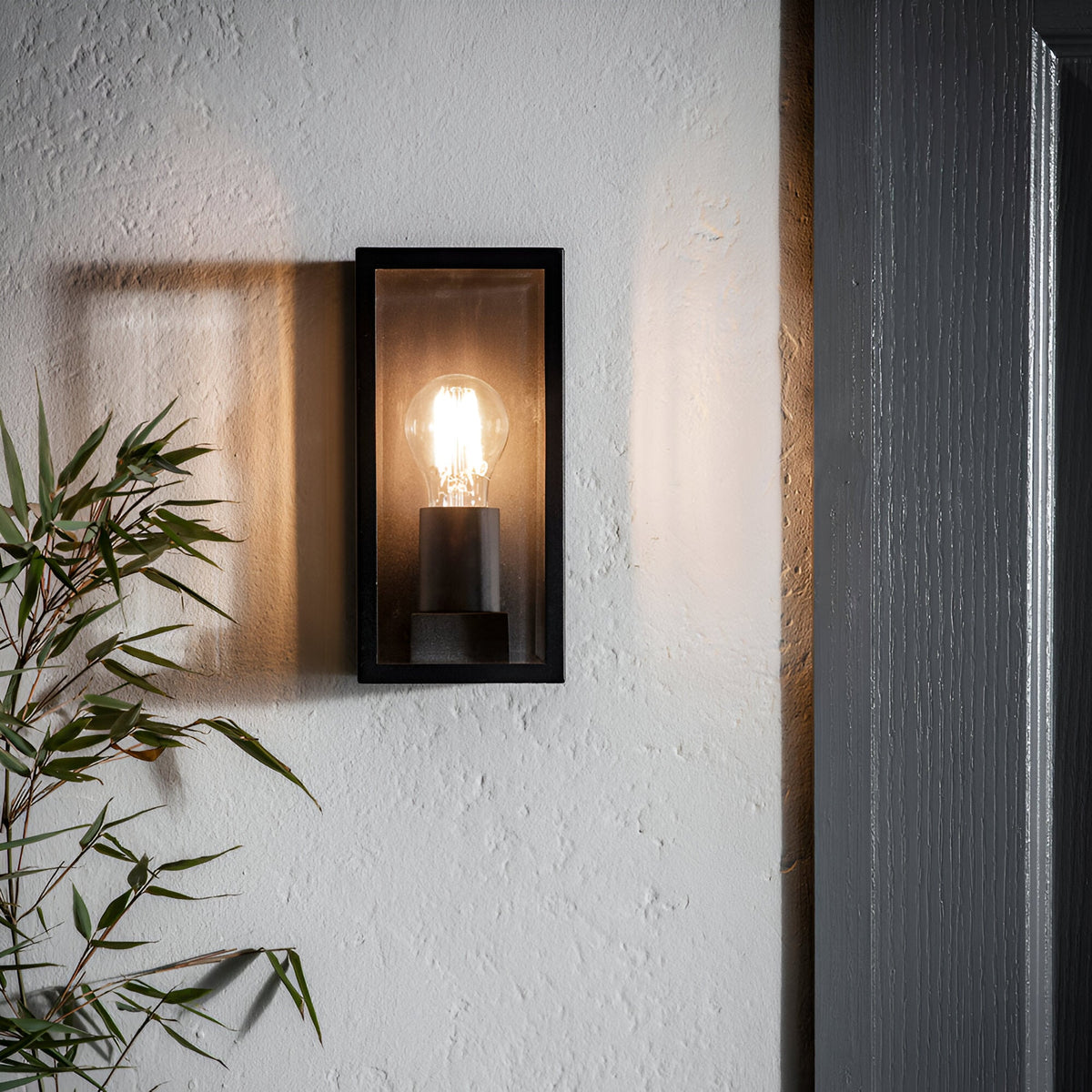 Endon Castwick box lantern wall light in matt black with bevelled glass panels and lit E27 filament bulb, mounted on a textured wall beside a doorway.