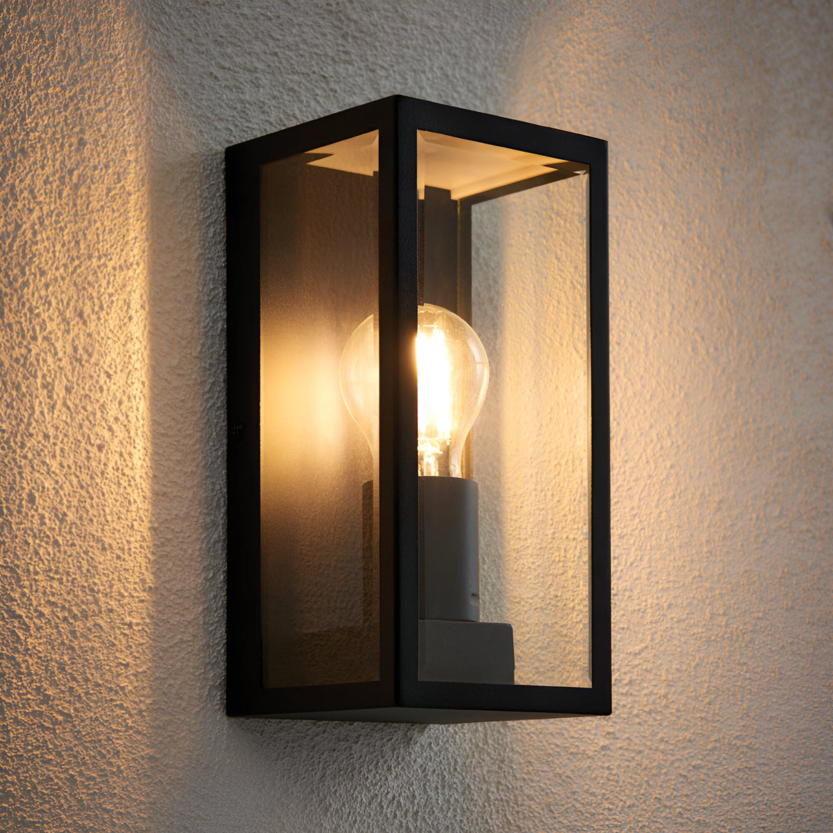 Endon Castwick box lantern wall light in matt black with clear bevelled glass panels and a single lit E27 filament bulb, mounted on a textured wall.