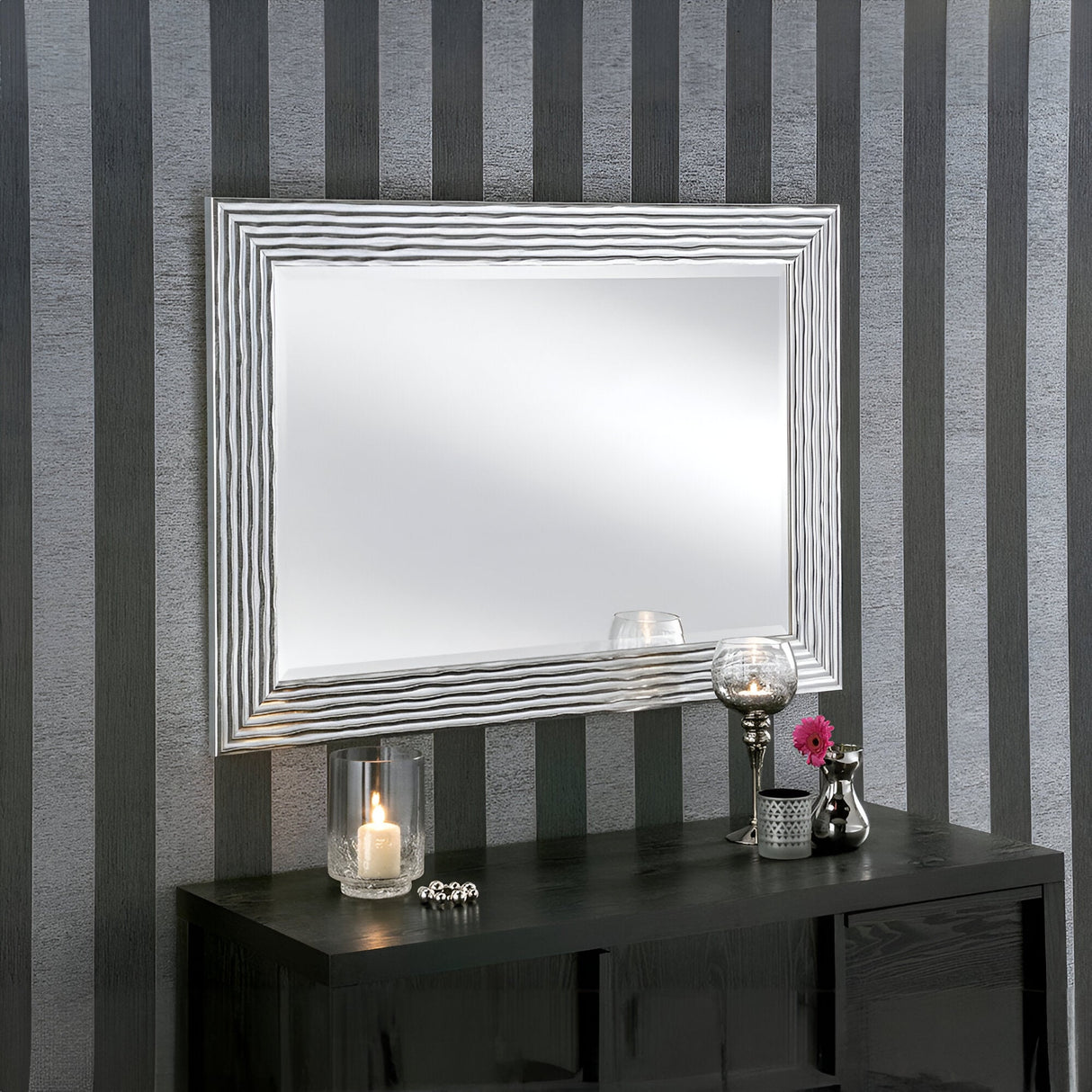 The Halo Rectangle Wall Mirror in silver with a textured frame is displayed on a striped gray wall above a sleek black console table. This modern design is enhanced by the table's arrangement of a lit candle, a small vase with pink flowers, a clear decorative orb, and a silver fruit bowl.