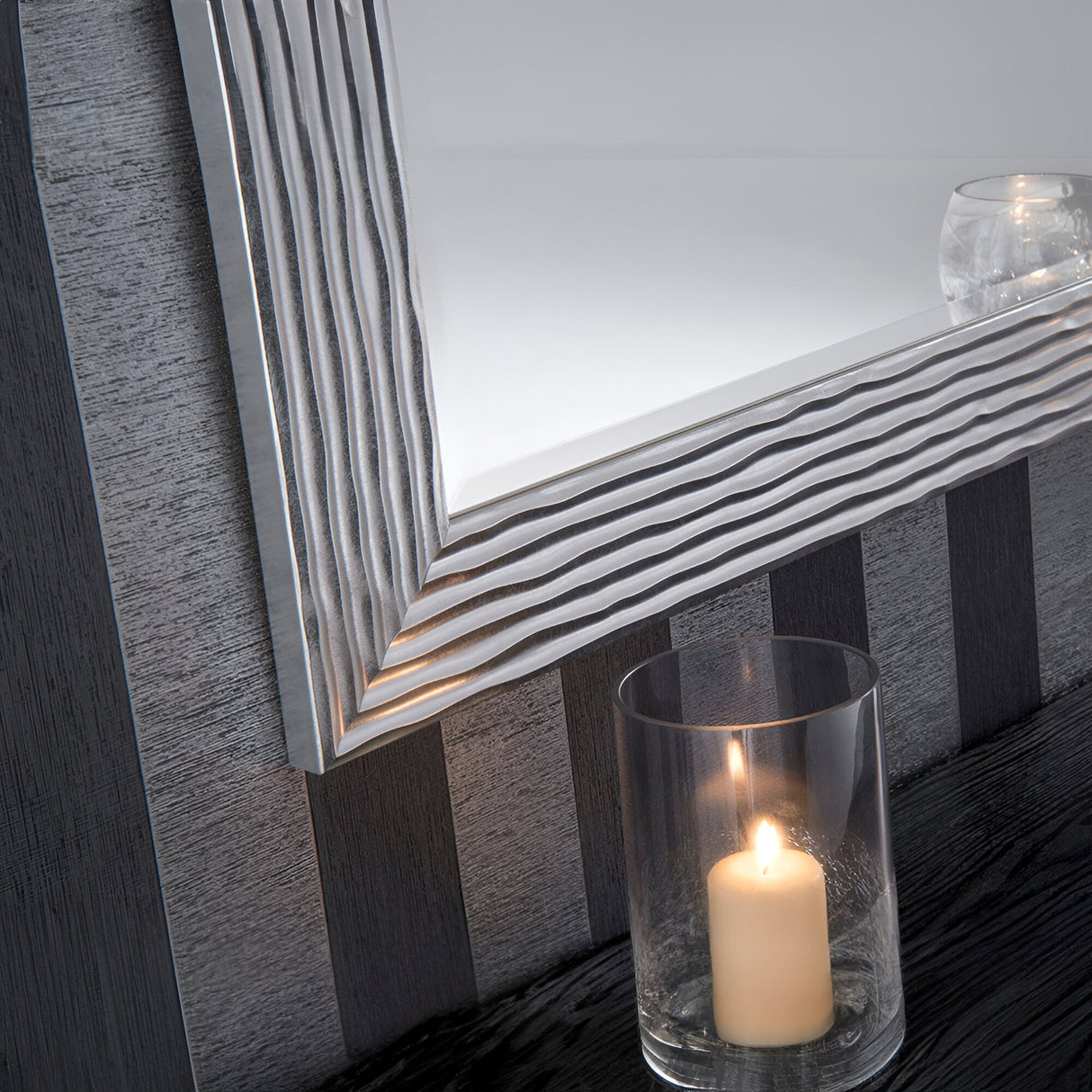 The Halo Rectangle Wall Mirror in silver, featuring a textured frame, is mounted on a striped wall. Beneath it, a lit candle rests gracefully in a clear glass holder on a dark surface. An additional small glass object is visible in the mirror's reflection, enhancing its modern design appeal.