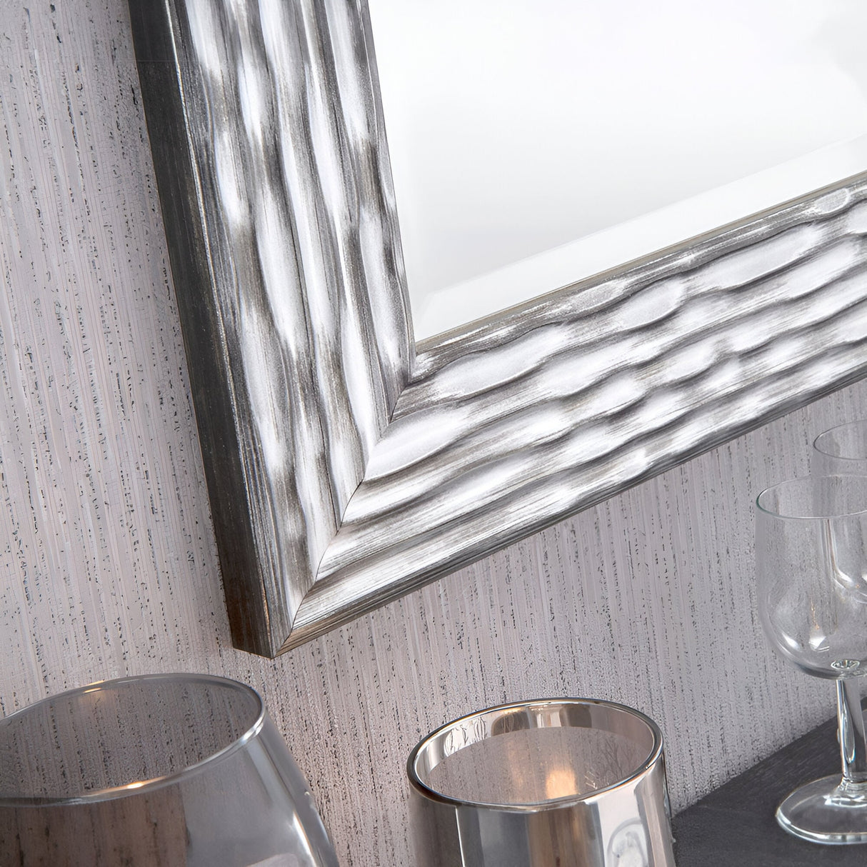 A Halo Rectangle Wall Mirror - Black infuses a space with modern sophistication through its silver, textured wave pattern. Installed on a light-colored, vertically striped wall, it captures the reflection of candle holders and empty wine glasses resting on a sleek dark surface below.