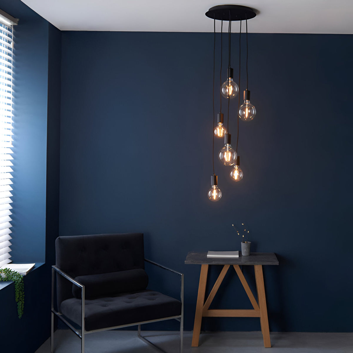 Marlowe 6 Light Pendant in matt black metal, six exposed bulbs on staggered fabric flex drops from a round ceiling rose, shown above a dark living room setting.