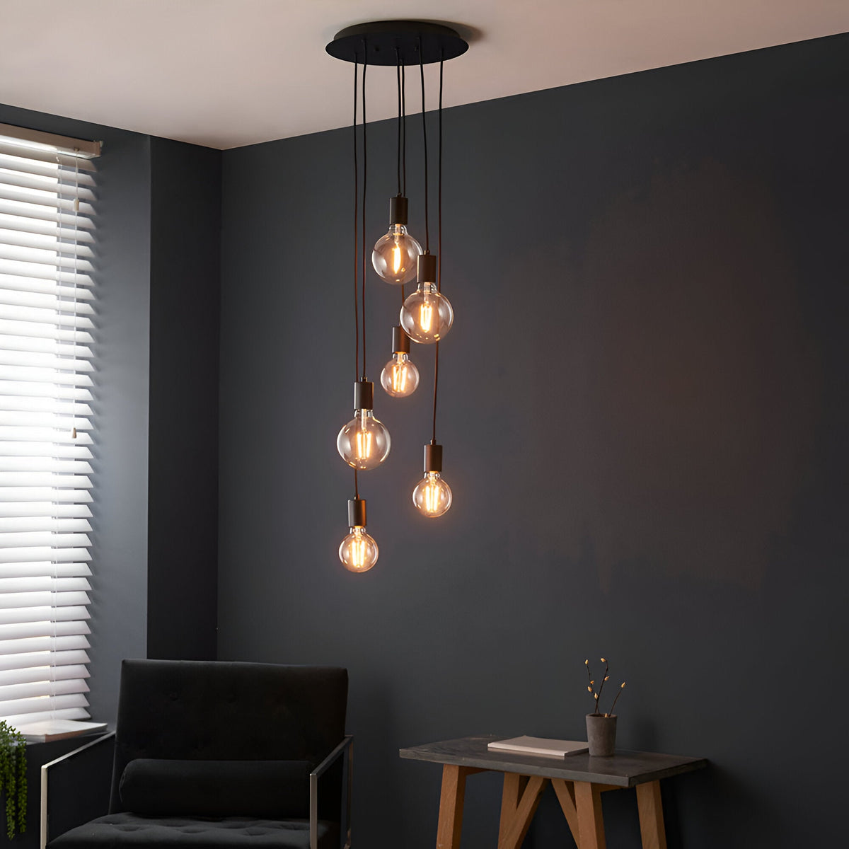 Marlowe 6 Light Pendant in matt black metal with exposed globe bulbs on staggered fabric flex drops, shown above a dark living room armchair and side table.