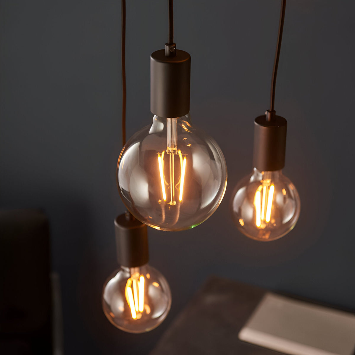 Marlowe 6 Light Pendant in matt black metal, showing three exposed E27 filament bulbs on staggered black fabric flex drops against a dark wall.