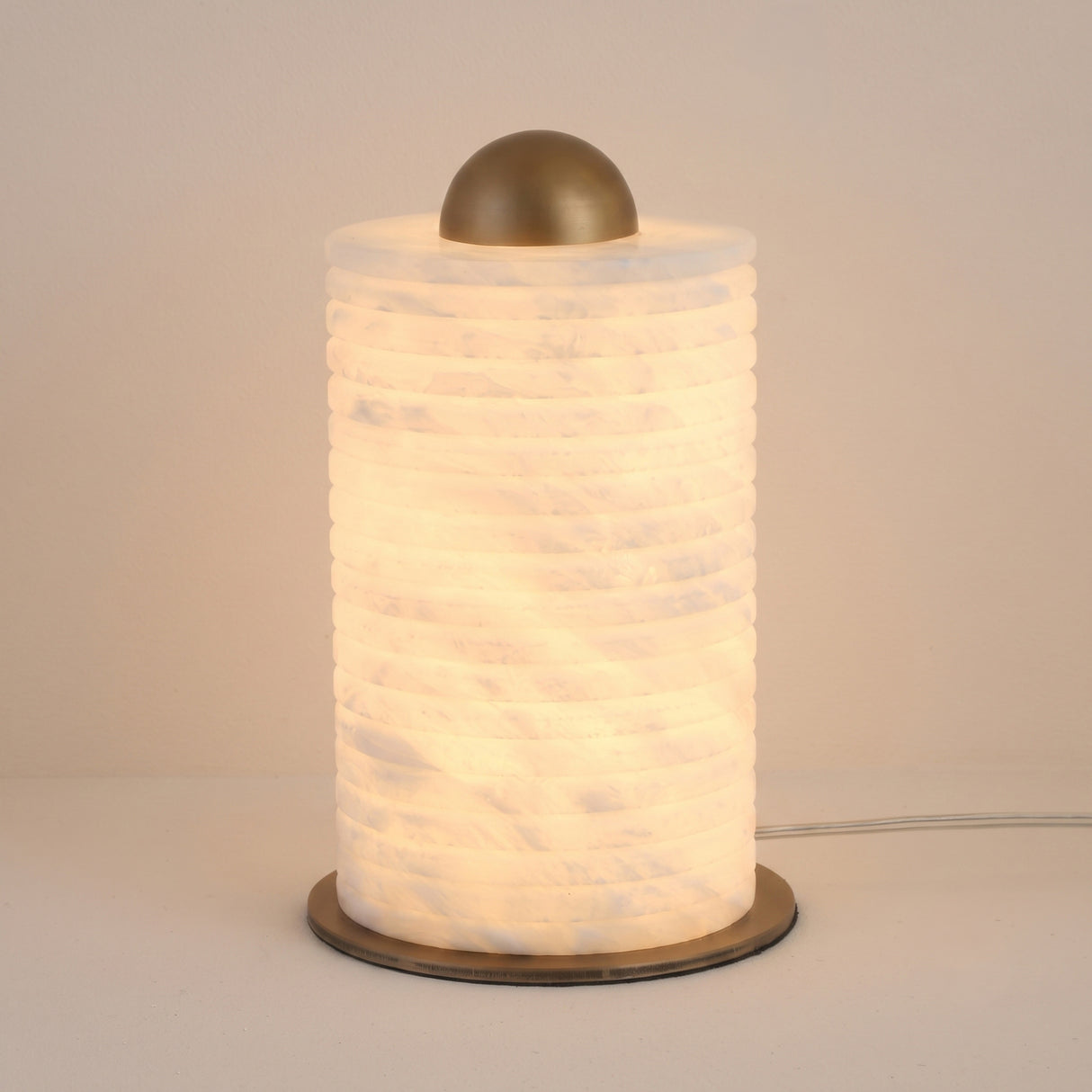 Aurelior Small LED table lamp with ribbed cylindrical alabaster body glowing warm white, topped by a brass dome cap and set on a slim brass base disc, shown against a plain light background.