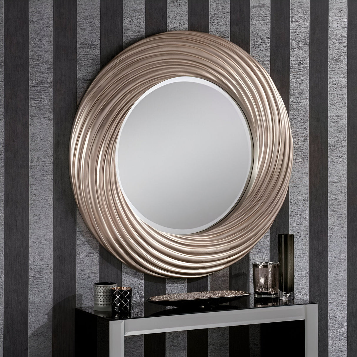 The Mirage Circle Wall Mirror Silver - 92cm, with its intricately textured metallic frame, hangs beautifully against a wall decorated in black and silver vertical stripes. Below it, the sleek black console table enhances the sophisticated home decor, with candles and a tray adding an extra touch of charm to the ensemble.