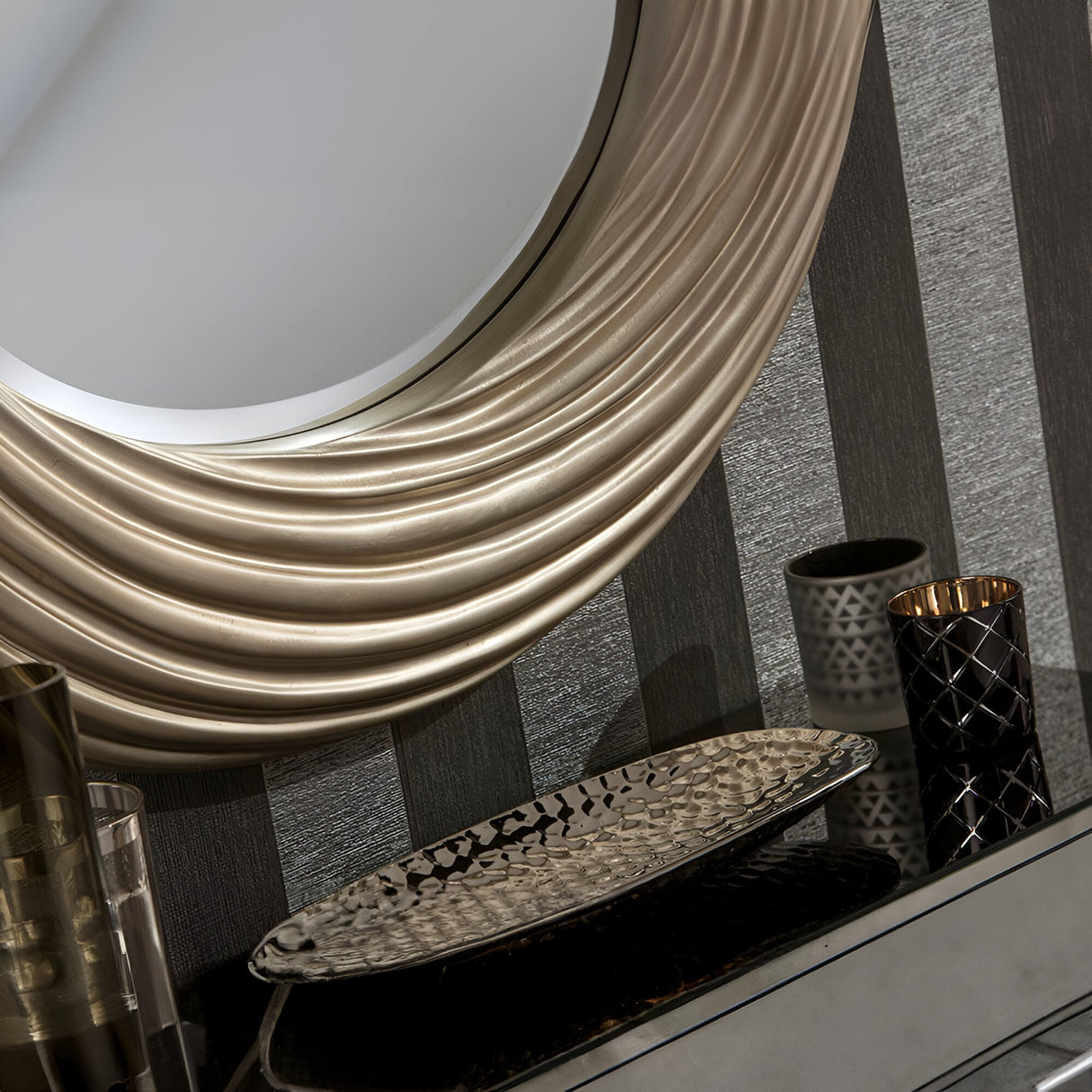 A decorative vanity setup featuring the Mirage Circle Wall Mirror Silver - 92cm with a textured silver frame. Below, elegant vases and a patterned tray rest on a reflective surface, enhancing the home decor against a chic striped wall.