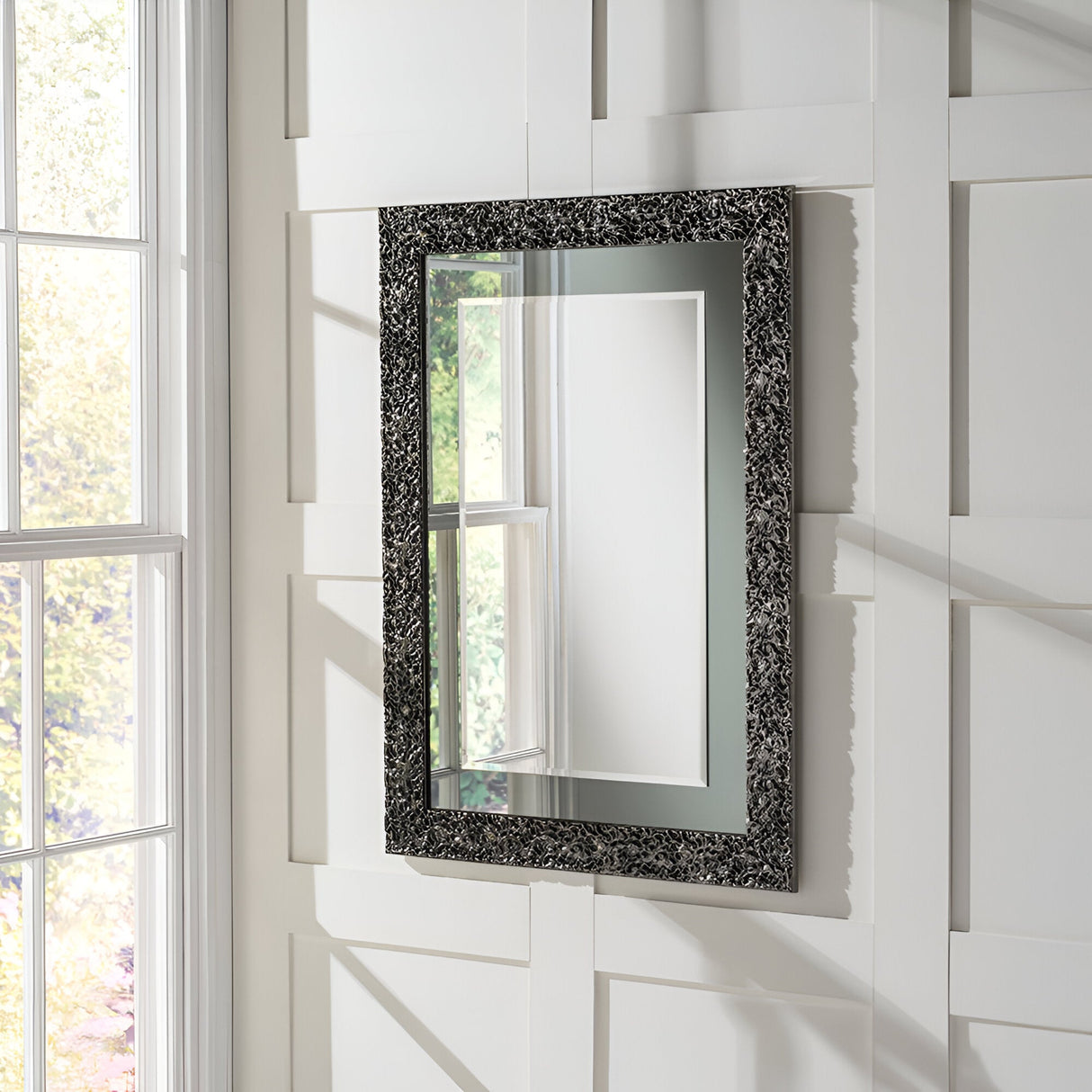 The Illume Rectangle Wall Mirror, featuring a grey lacquered frame with an intricate design, hangs on a light-colored paneled wall, bringing a modern sophistication to the home decor. Positioned next to a large window on the left, it reflects the natural light and outdoor scenery beautifully in its sleek finish.