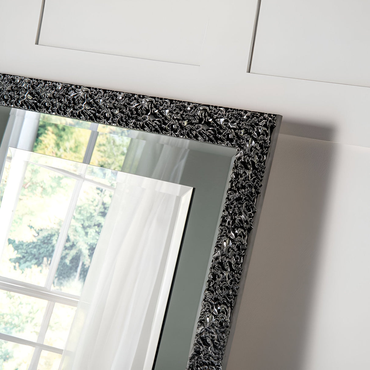 The Illume Rectangle Wall Mirror - Grey Lacquered features an intricately designed black and silver frame that exudes modern sophistication, perfectly reflecting a window view of verdant trees and white curtains. Its lacquered finish adds elegance as it leans against a white paneled wall, enhancing the charm of your home decor.