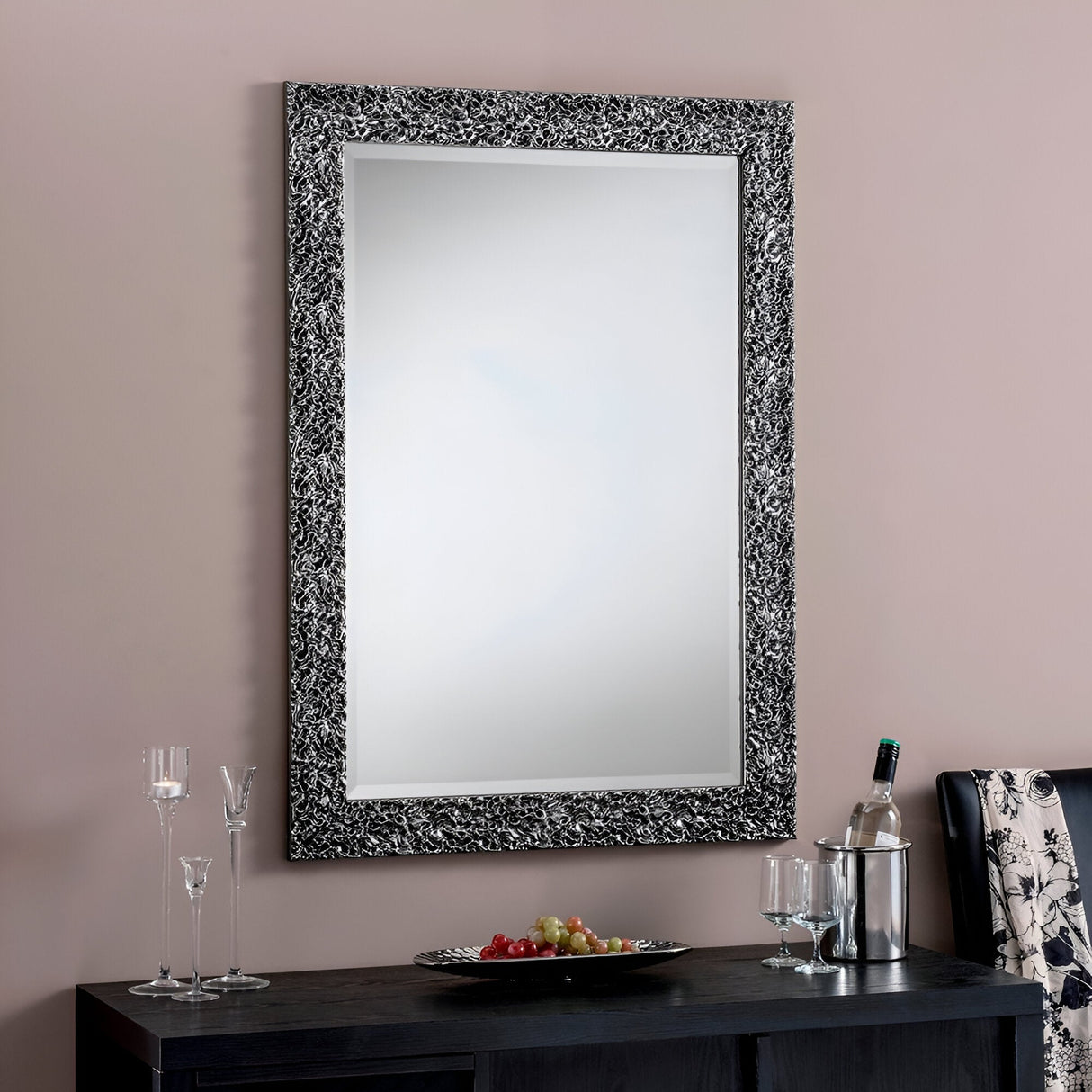 The Illume Rectangle Wall Mirror - Grey, with its decorative black and grey frame, hangs on a pink wall. Below it, a sleek black console table complements the modern decor by displaying a platter of grapes, wine glasses, and a wine bottle chilling in an ice bucket.