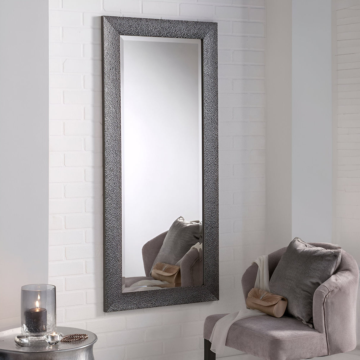 The Eclipso Rectangle Wall Mirror - Grey, featuring its textured metal frame, radiates modern elegance on a white brick wall. Positioned in front is a plush gray chair with a pillow, paired with a small round table that holds a black candle to complete the sophisticated ambiance.