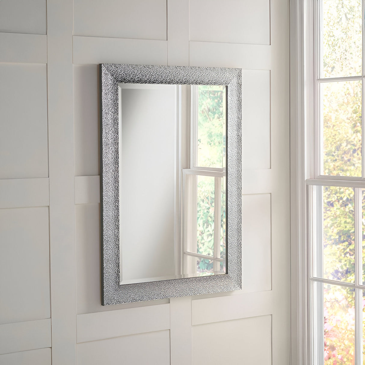 The Eclipso Rectangle Wall Mirror - Silver, featuring its textured silver frame, is gracefully mounted on a white paneled wall. It wonderfully reflects a portion of a window with views of lush trees and a garden, enhancing the allure of the modern interior.