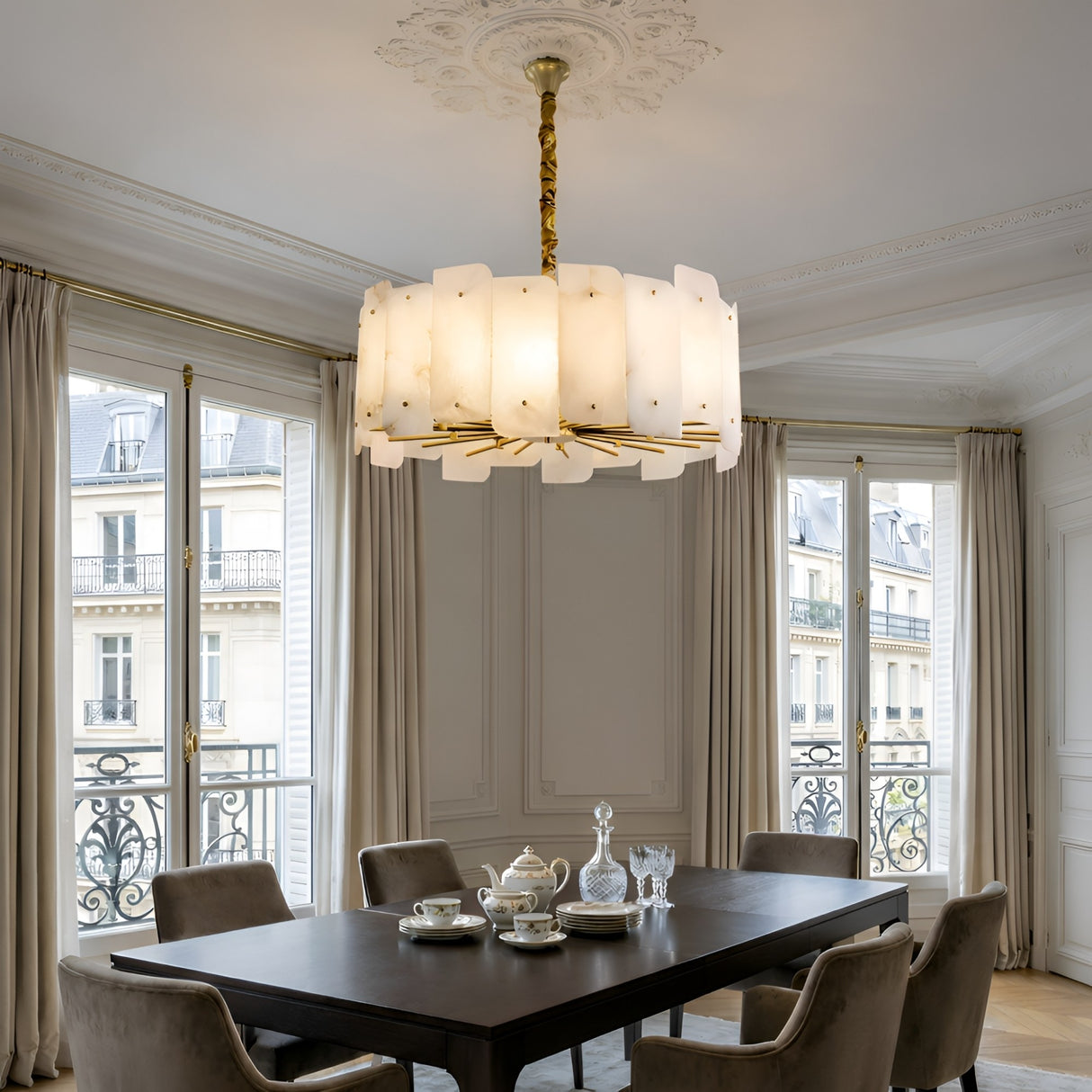 The Caldrin 6 Light Tiered Panel Alabaster Chandelier in Soft White & Brass hangs over a dark dining table with tea service in an elegant room featuring tall windows, cream curtains, ornate moulding, and views of Parisian-style buildings.