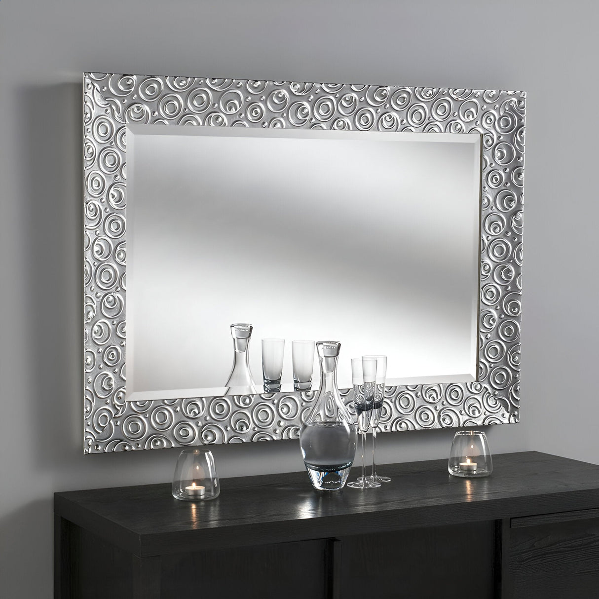 The Astral Rectangle Wall Mirror - Silver, with its ornate frame featuring concentric circles, hangs on a gray wall. Below it, a dark wooden console displays several glass bottles and teardrop-shaped candle holders. The reflections on the mirror's surface shimmer, adding depth to the elegant setting.