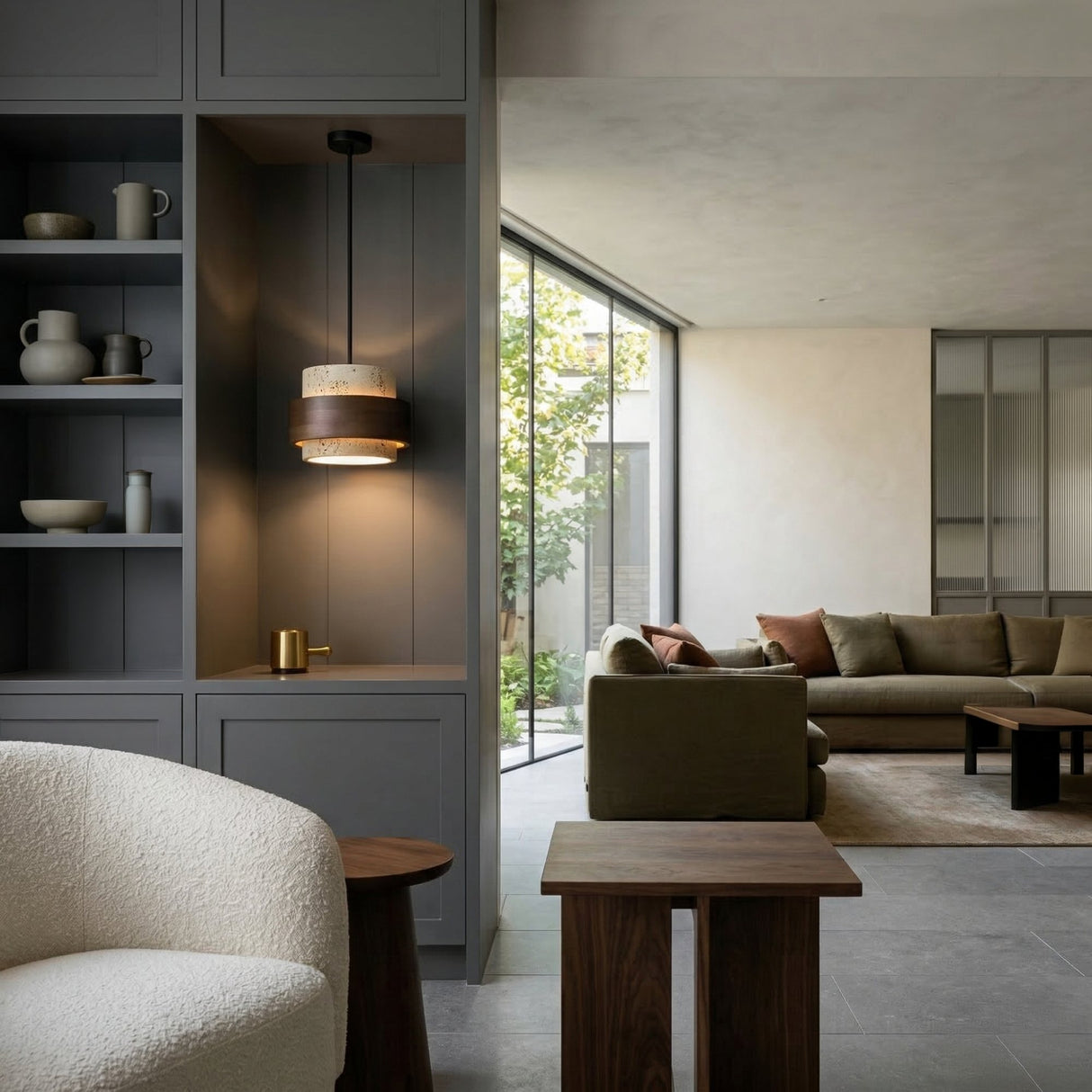Modern living room with gray built-ins, a white textured armchair, wood tables, a cozy green sofa, and bright garden-facing windows—illuminated by the Virelli LED Drum Travertine Single Pendant Light in Walnut Brown & Light Beige overhead.