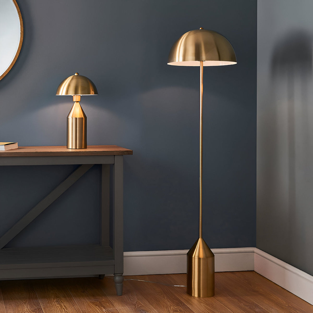 Castford Table Lamp in antique brass with a domed metal shade on a wooden console, beside a matching antique brass floor lamp in a living room.
