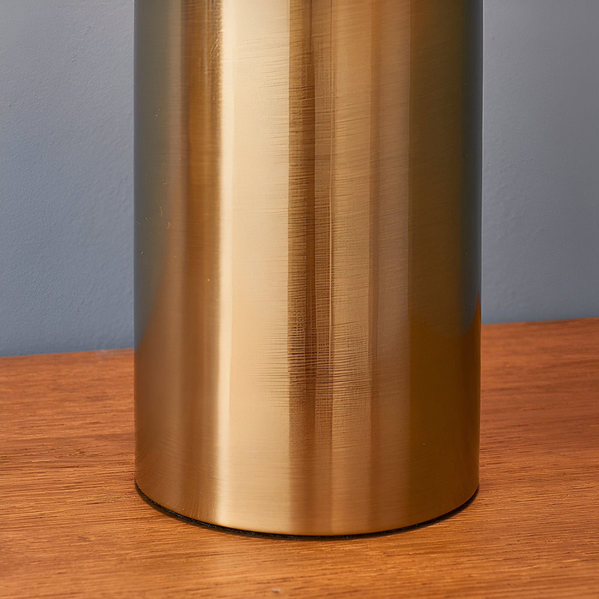 Close-up of the Castford Table Lamp's cylindrical base in antique brass with a brushed metal finish, shown on a wood surface against a grey wall.