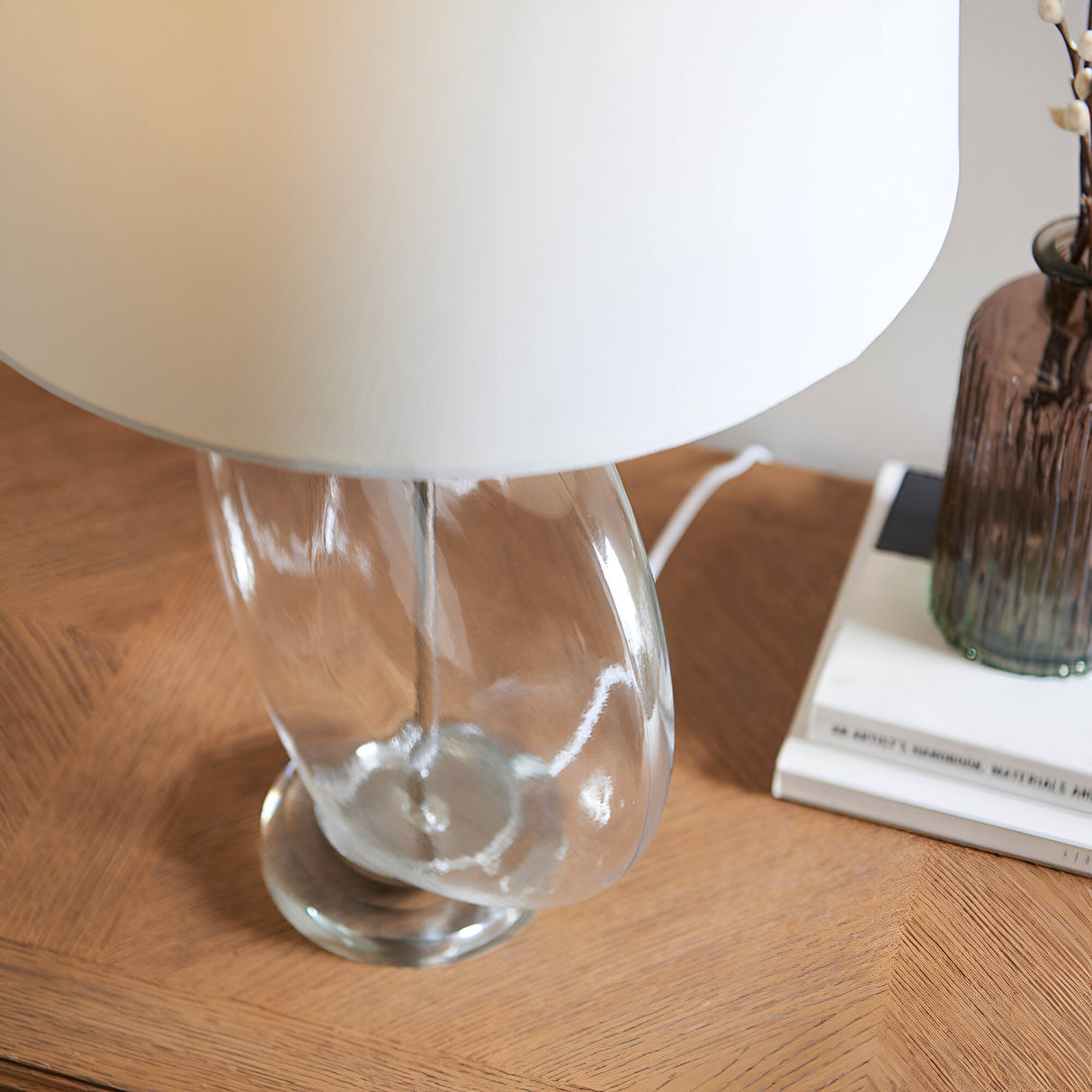 Close-up of the Endon Bramleigh table lamp's clear glass ellipsoid base and white shade on a wooden console beside books and a brown glass vase.