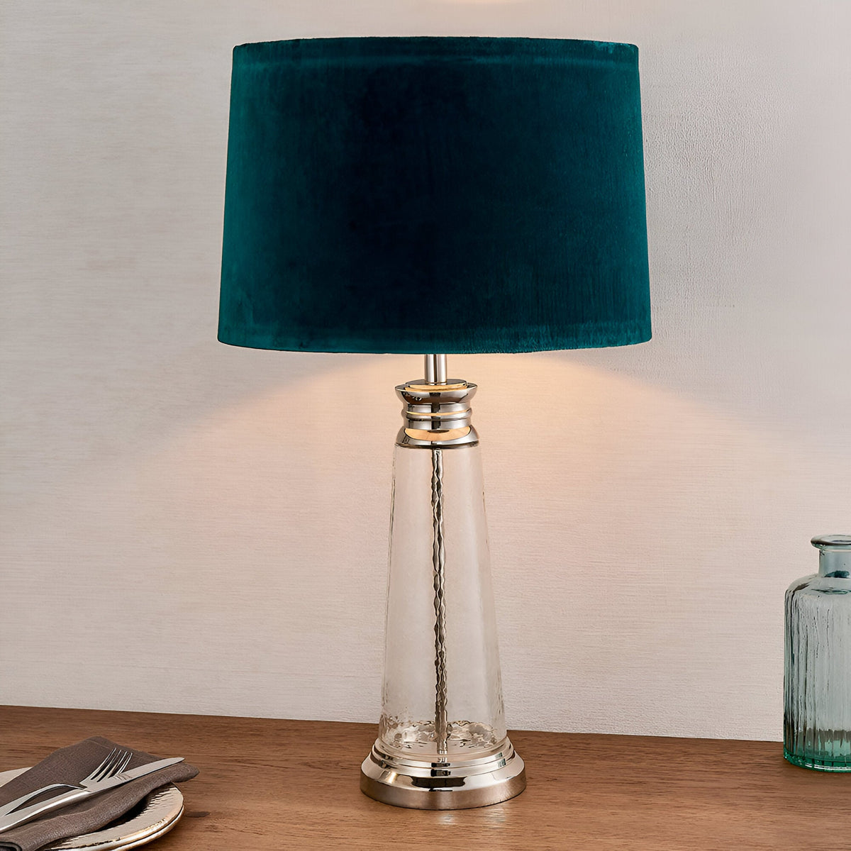 Endon Danehurst table lamp with clear hammered glass base, nickel metalwork and teal velvet tapered shade, lit on a wooden sideboard.