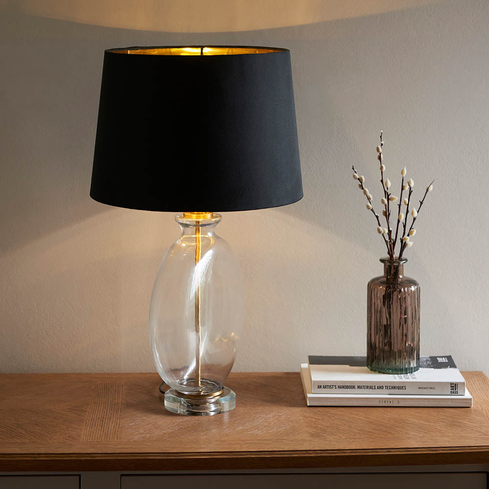 Endon Bramleigh table lamp with clear glass ellipsoid base and black tapered shade with brass interior, on a wooden console beside books and a vase.