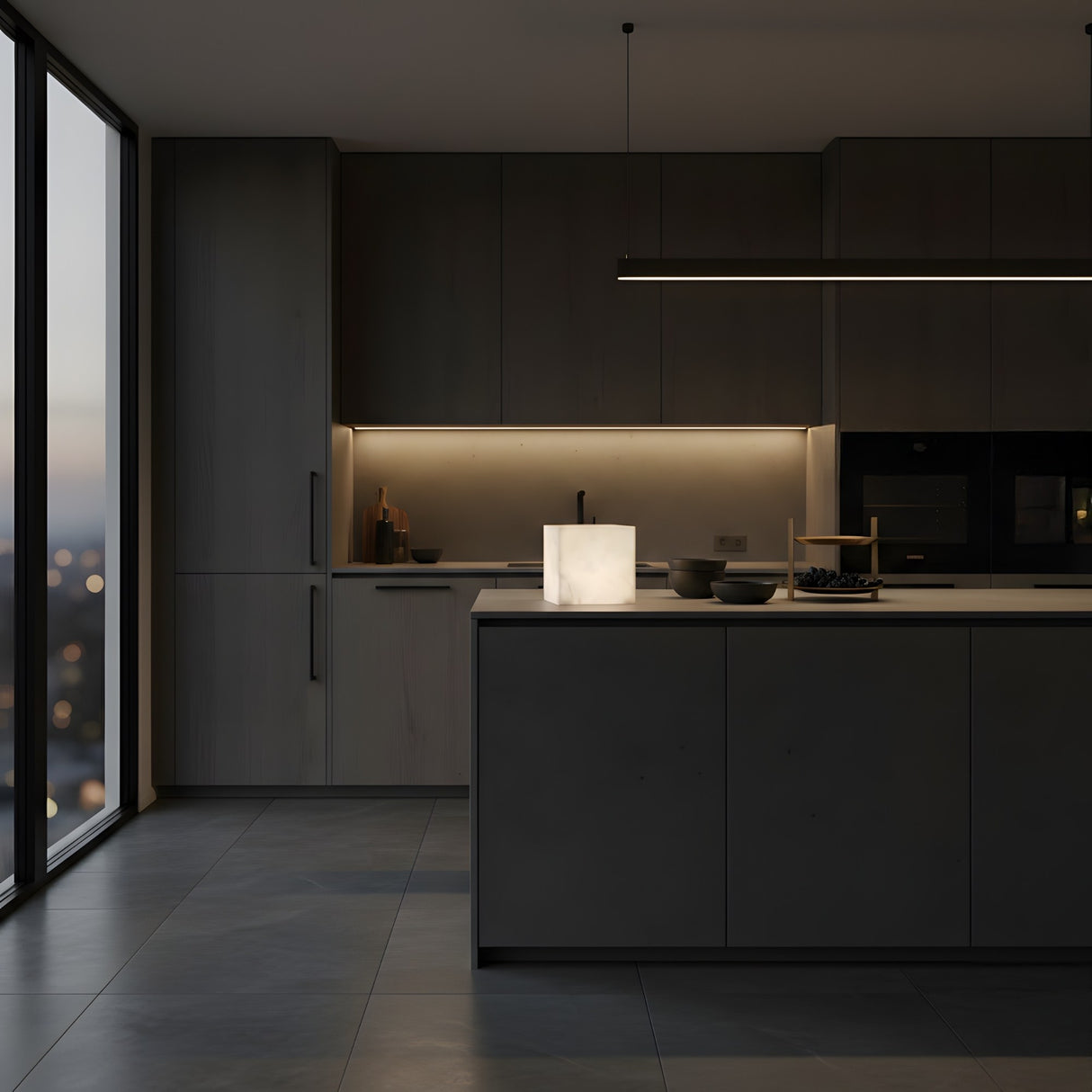 A modern kitchen with dark cabinets, an island, built-in appliances, and soft under-cabinet lighting. A glowing Cubara LED Small Cube Alabaster Table Lamp – Soft White sits on the island as large windows showcase an evening cityscape.