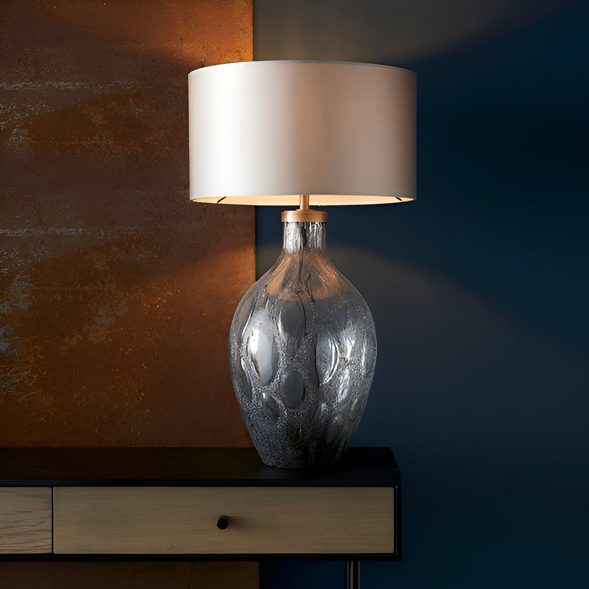 Endon Brenstead table lamp with charcoal textured artisan glass base, antique brass fitting and cream drum shade on a wooden sideboard.