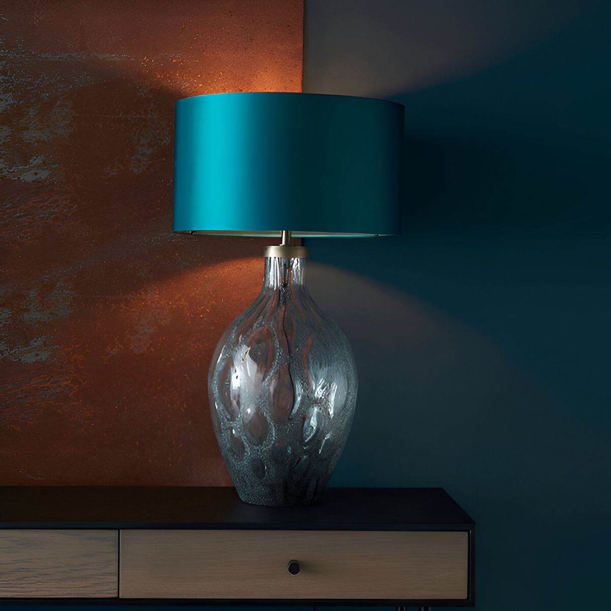 Endon Brenstead table lamp with charcoal-tinted textured glass base and antique brass detail, topped with a teal satin drum shade on a wooden sideboard.