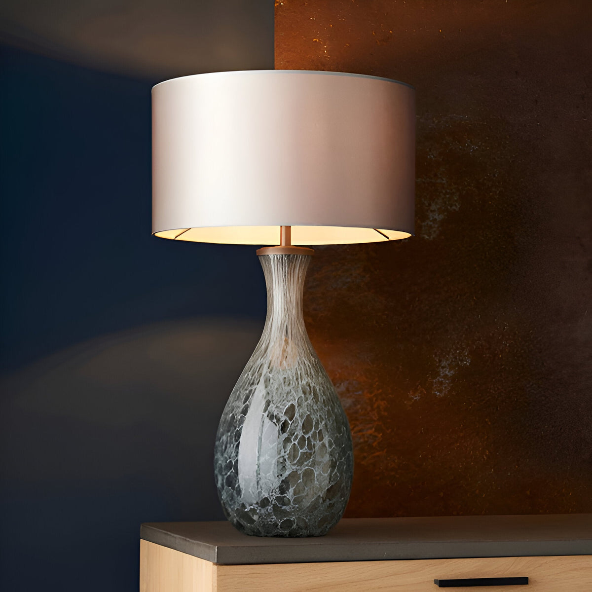 Carldale Table Lamp in Bronze with grey, cloud-effect glass hourglass base and matching fabric drum shade, shown lit against a dark blue and rust-toned wall.