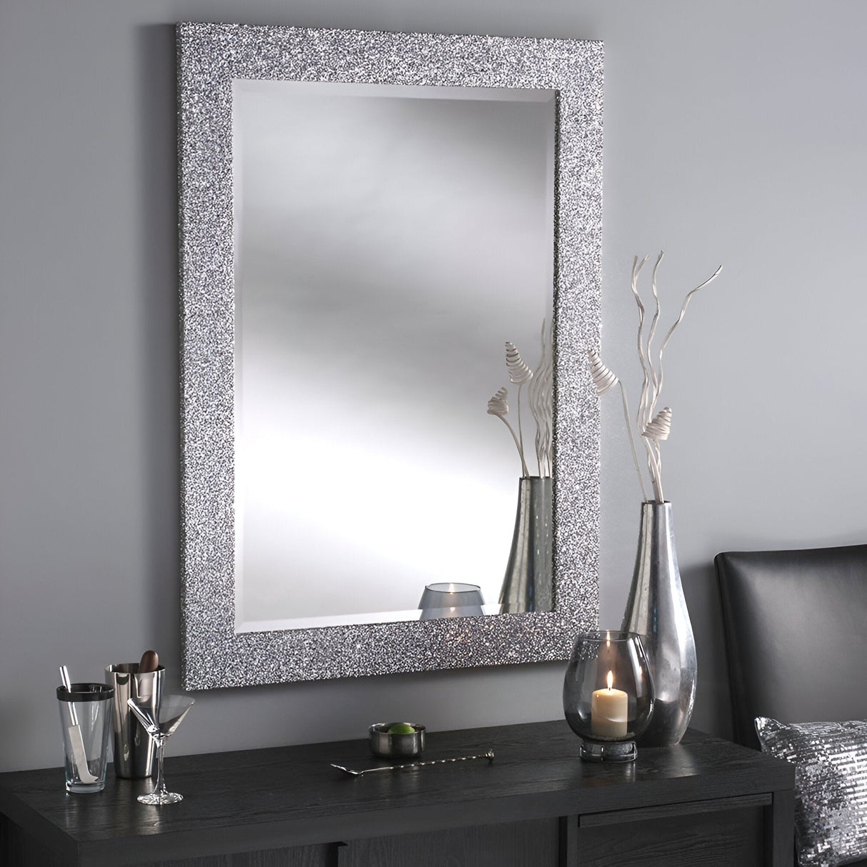 The Fusion Rectangle Wall Mirror in silver hangs elegantly on a gray wall. Below it, a dark dresser displays decorative items: a vase with white branches, a candle in a glass holder, and a silver cup containing small tools. Part of the bed with its gray headboard is also visible.