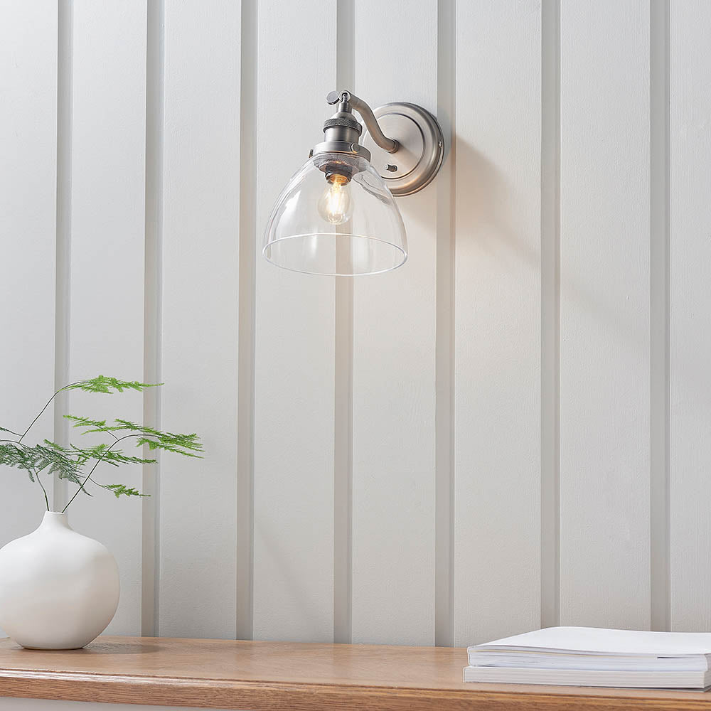 Silver Wentworth wall light with clear glass dome shade and adjustable knuckle arm, shown lit above a wooden shelf with a vase and plant.