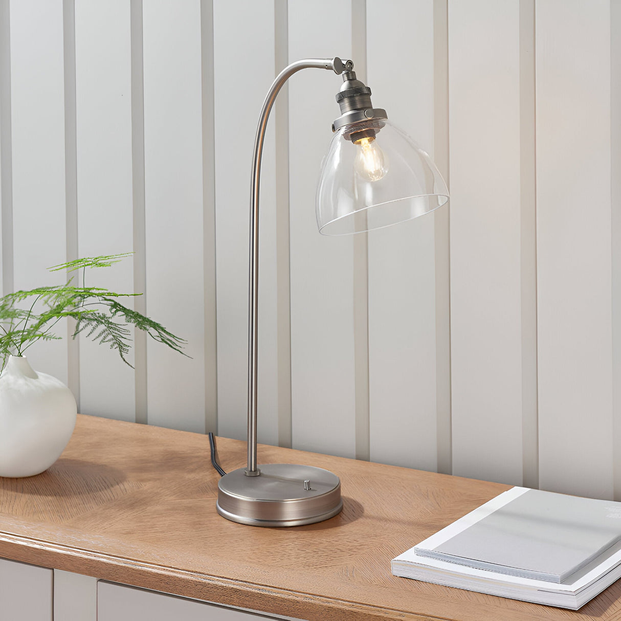 Endon Wentworth industrial table lamp in silver with arched stem, clear glass dome shade and toggle switch, on a wooden console beside a fern.