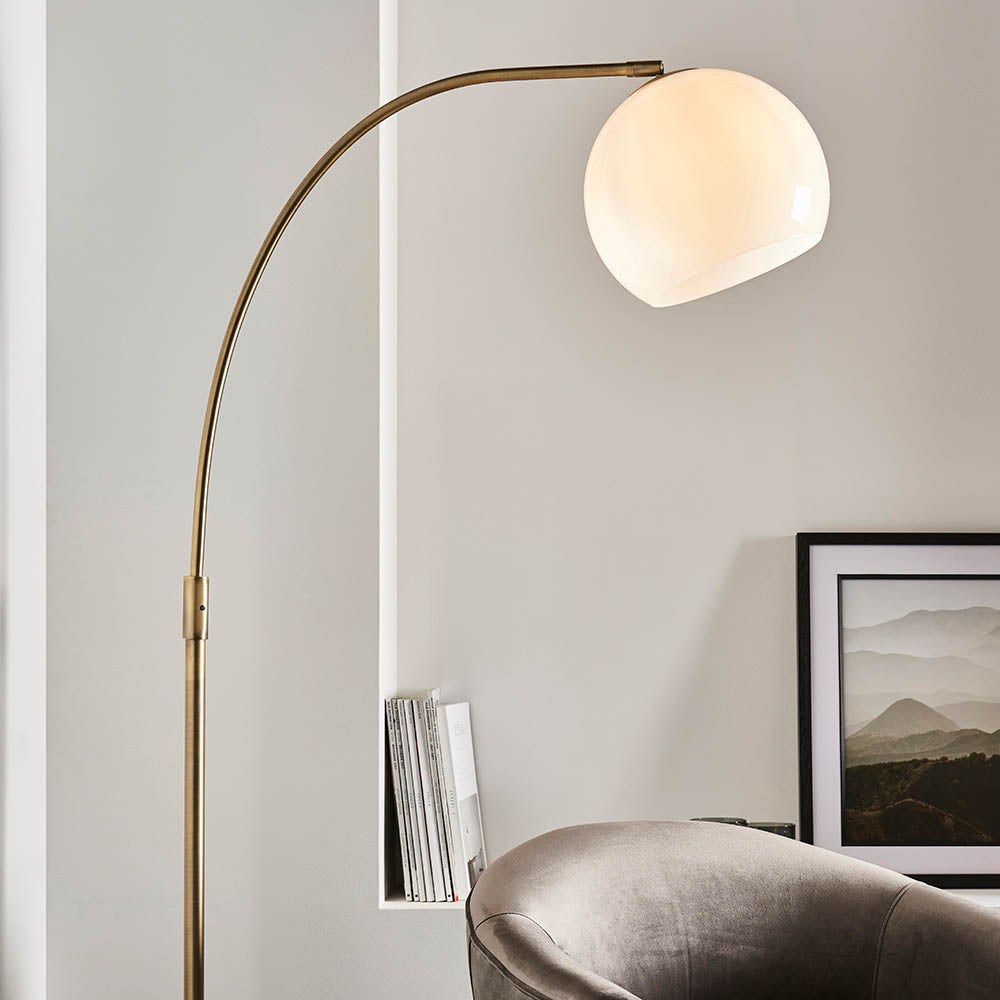 Sinclair Floor Lamp with curved brass stem and round white glass globe shade, shown in a living room beside an armchair and framed print.