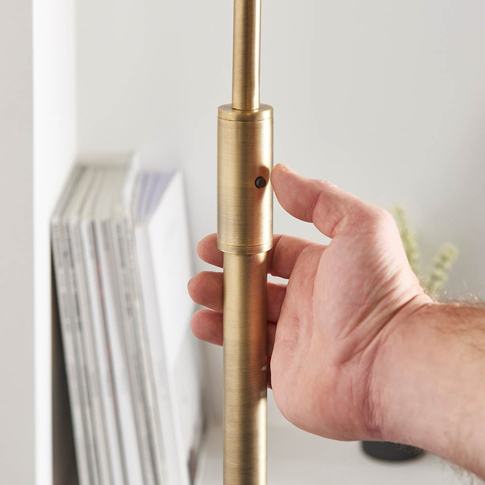 Close-up of a hand adjusting the brass telescopic stem of the Sinclair Floor Lamp near a shelf of books.