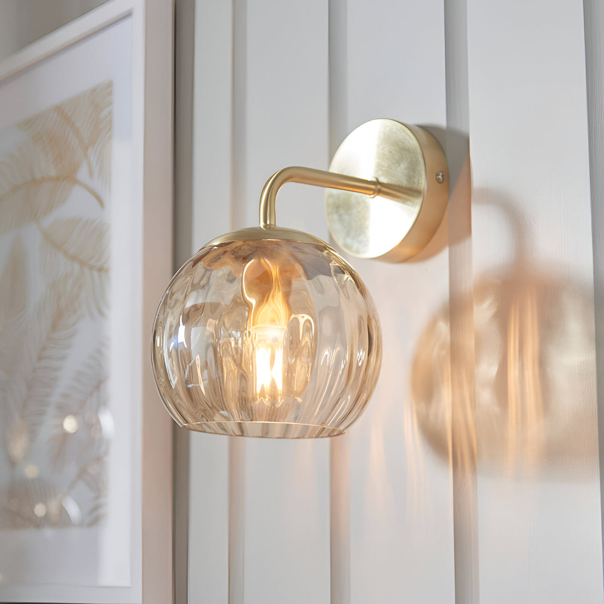Endon Draycott wall light with champagne dimpled glass globe shade on a satin brass arm and round backplate, illuminated beside a framed print.