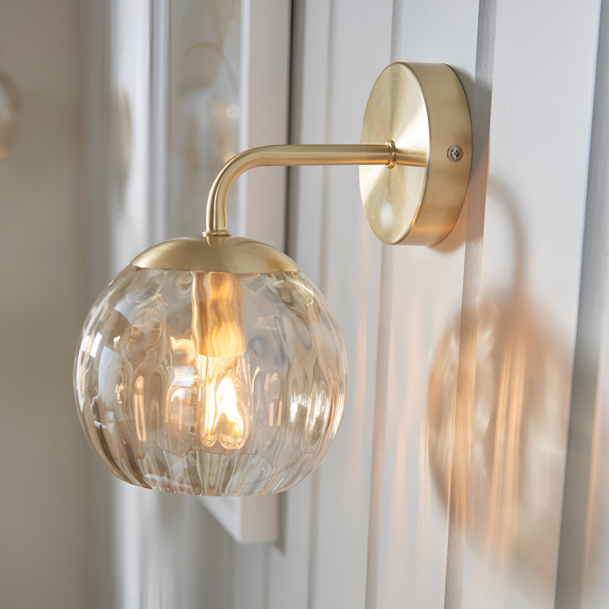 Endon Draycott wall light with champagne dimpled glass globe shade on a satin brass arm and round backplate, mounted on white panelled wall.