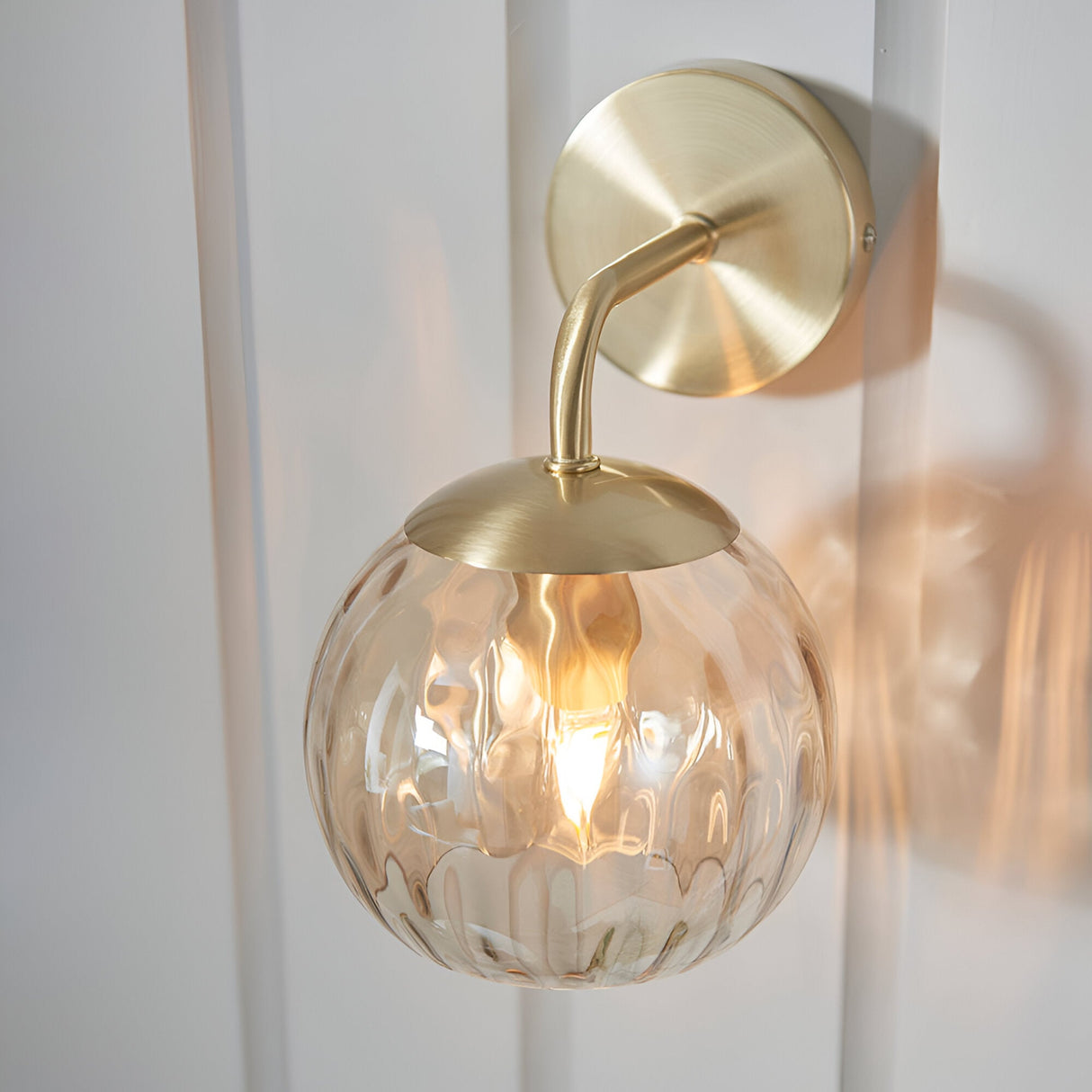 Endon Draycott wall light with a champagne lustre dimpled glass globe shade on a satin brass arm and round backplate, mounted on a white panelled wall.