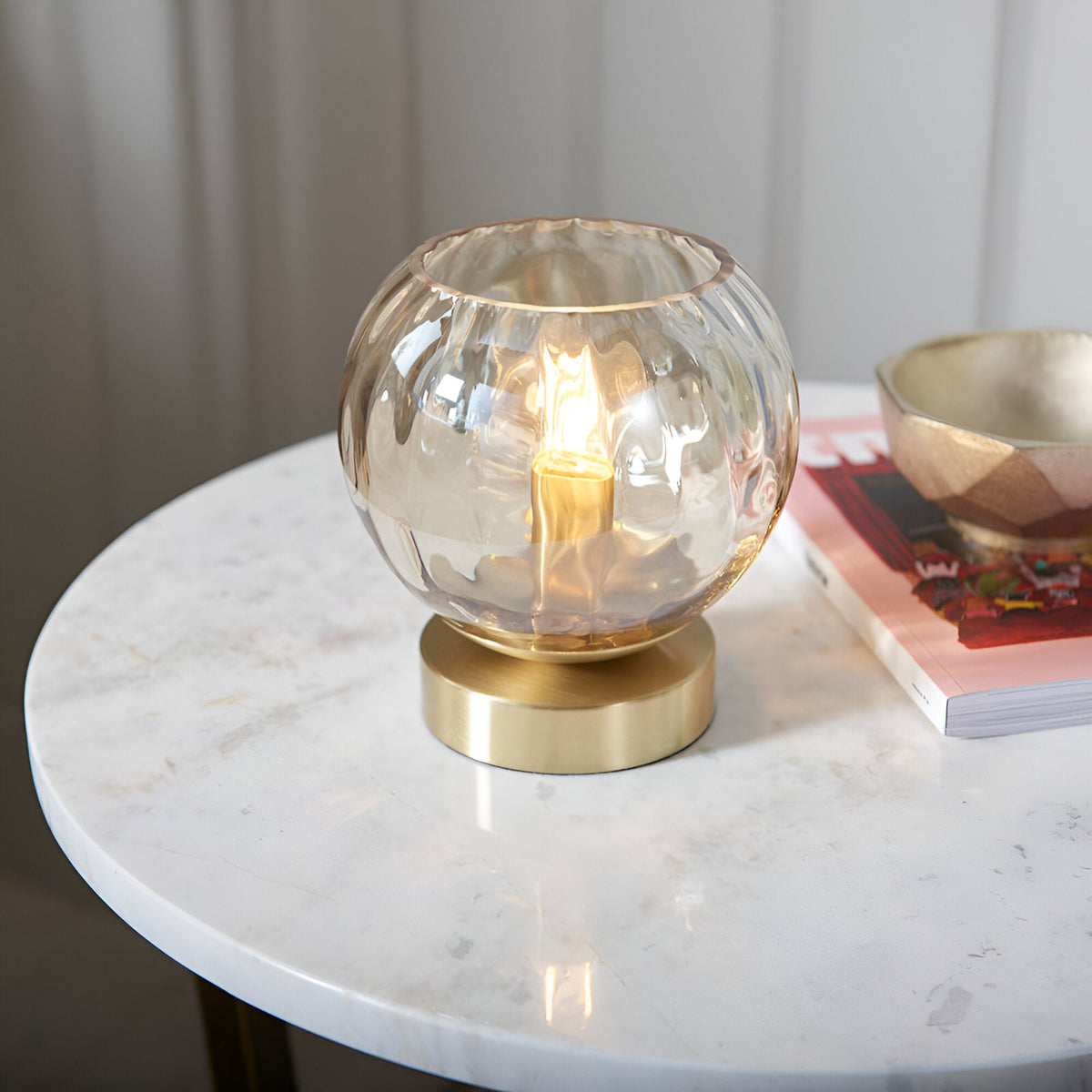 Draycott Dimpled Glass Table Lamp in champagne glass with a satin gold base, lit on a marble side table beside a magazine and dish.